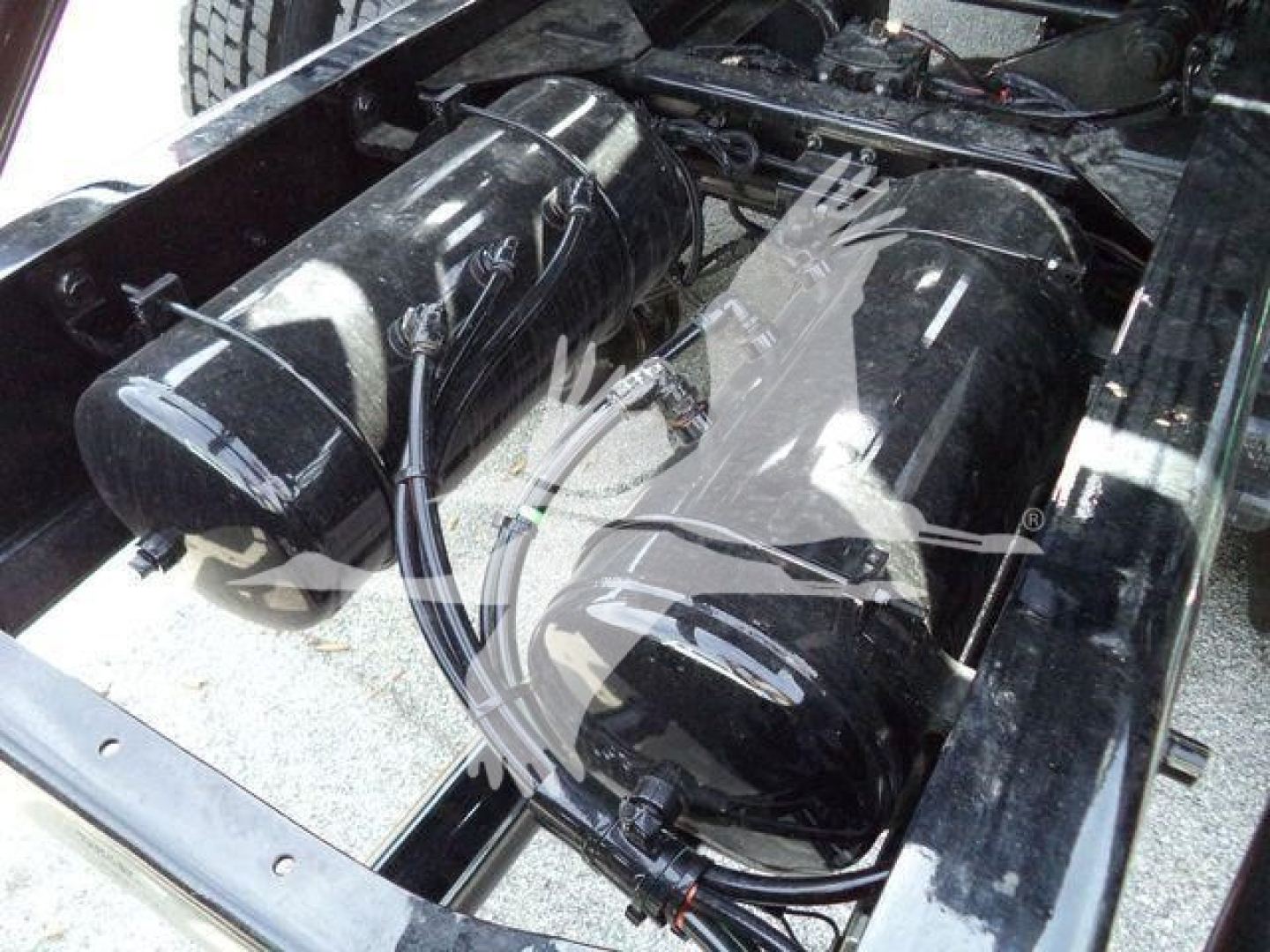 Dual cylindrical fuel tanks with hoses in a truck bed area, mounted between chassis rails; black glossy surface with reflection.
