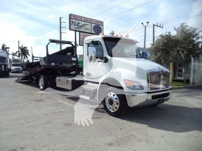 Truck bay showing a white 2025 Kenworth T280 rollback tow truck with a flatbed in a dealership lot, WKWT280XLP22 stock number visible.