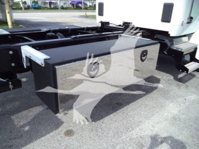 Kenworth truck chassis with a large rectangular black toolbox mounted on the side of the frame near the cab; visible metal brackets and steps; white cab in background.