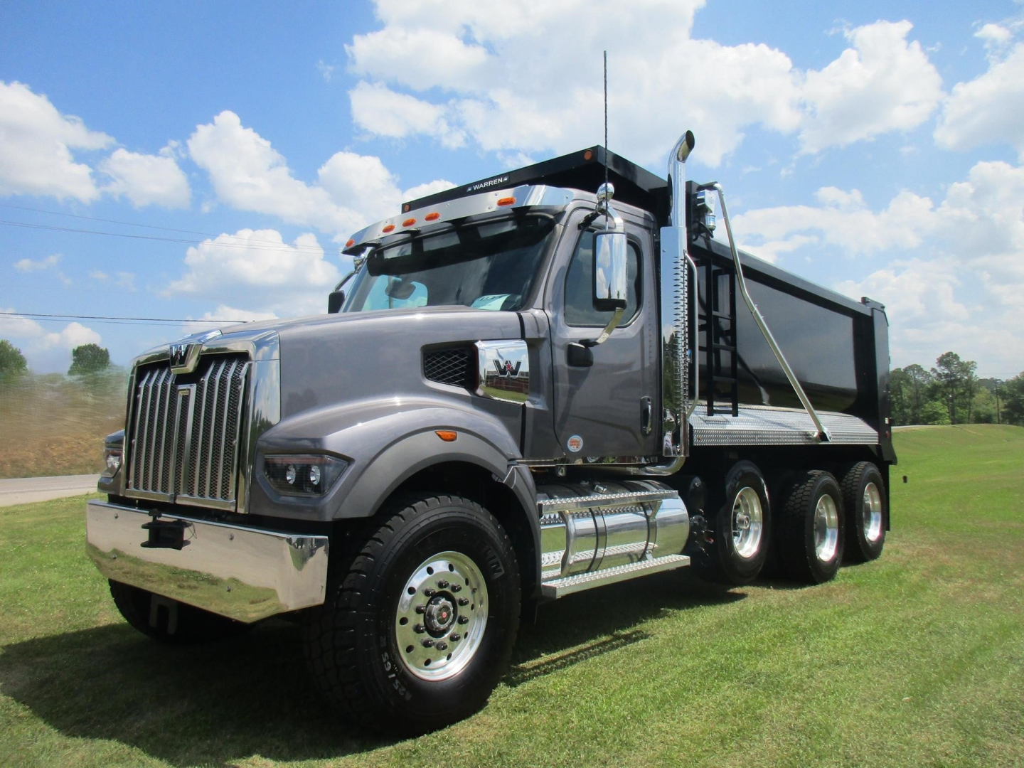 Truck image: 2026 Western Star 49X dump truck with gray cab, black grille, chrome bumper, and dump bed on a grassy field.