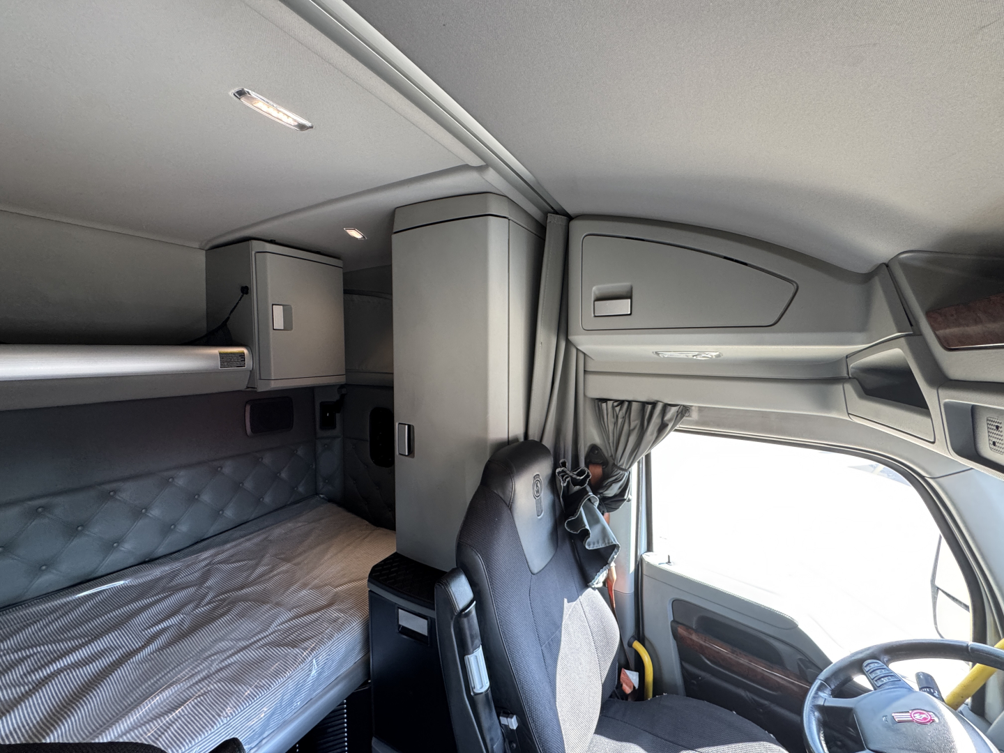Kenworth T680 cab interior showing sleeper berth with quilted padding, overhead storage, and driver seat; dashboard and windshield visible.