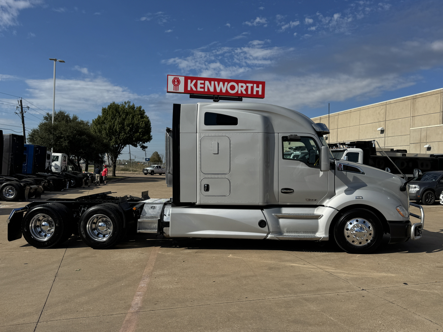 Kenworth T680 conventional sleeper tractor with silver exterior, side view showing cab, dual rear axles, Kenworth sign in background.