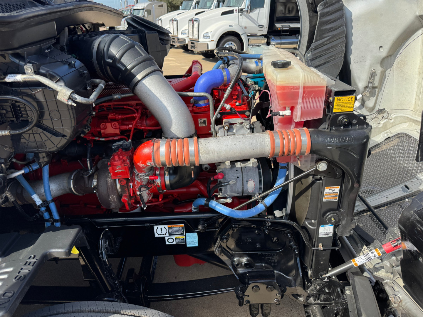 Kenworth T680 engine bay with red engine, blue and orange hoses, coolant reservoir, and intercooler piping visible.