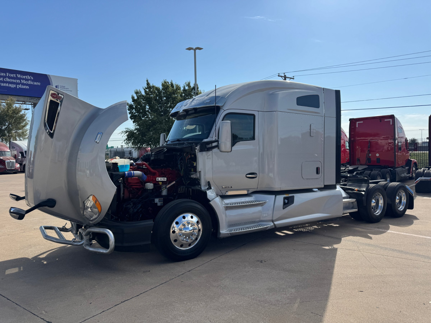 Kenworth T680 semi-truck with hood open showing engine bay; cab is light gray; chrome front wheel covers; visible side exhaust; no other visible text.