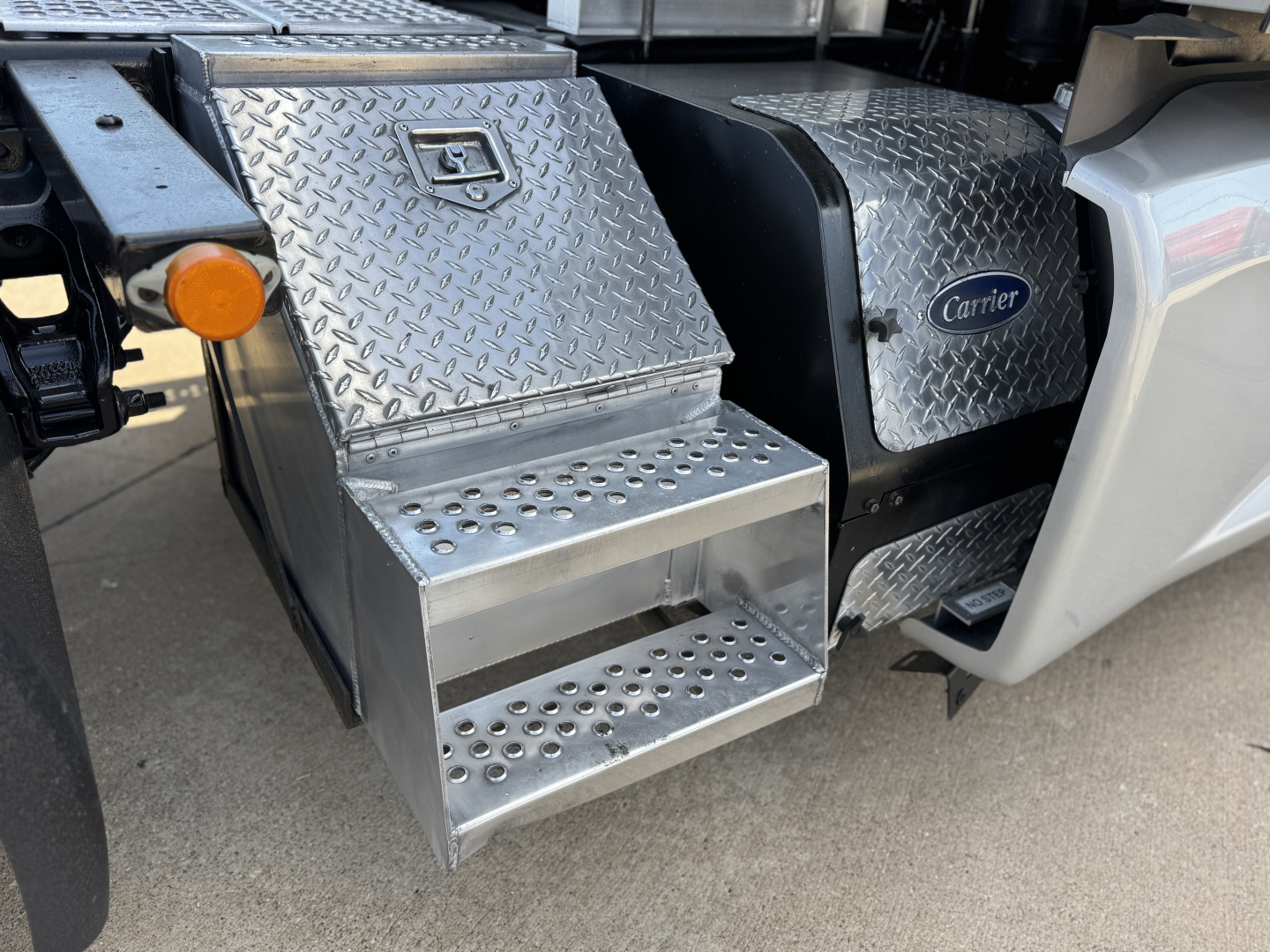 Kenworth T680 side step area with diamond plate toolbox and perforated metal step showing Carrier badge and chassis details.