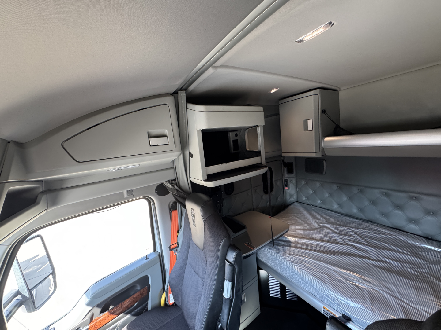 Kenworth T680 sleeper cab interior showing overhead storage shelf, bunk bed with mattress, and cockpit area with seats.