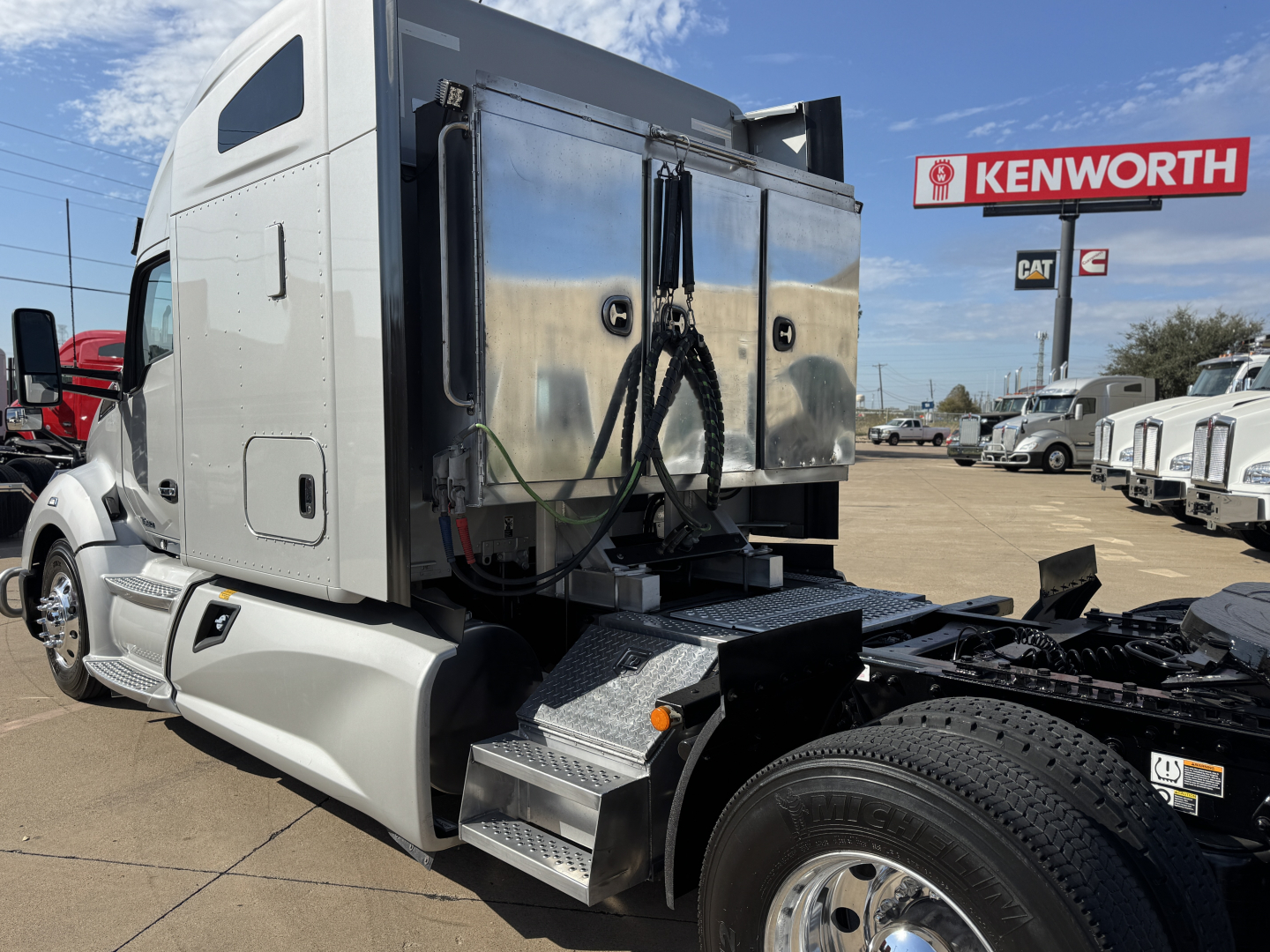 Kenworth T680 tractor cab side and fuel/auxiliary compartments with chrome steps and rear fender, adjacent Kenworth dealership sign.