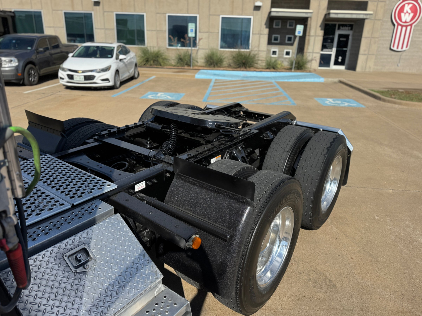 Kenworth T680 truck chassis view showing dual rear axles, fifth-wheel mount, and diamond-plate toolbox; front of a dealership building in background.