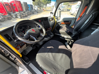 Kenworth T680 truck interior cab showing steering wheel with Kenworth emblem, dashboard, pedals, and driver seat; daylight through windshield.