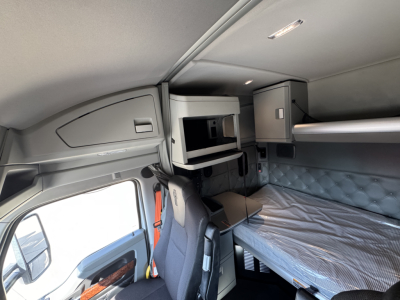 Kenworth T680 sleeper cab interior showing overhead storage shelf, bunk bed with mattress, and cockpit area with seats.