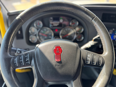 Kenworth T680 steering wheel and gauge cluster in cab; visible circular instrument dials behind steering wheel; Kenworth emblem center; dashboard controls on both sides.