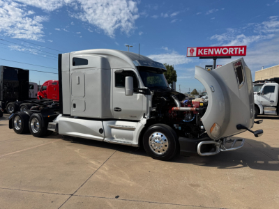 Kenworth T680 tractor with hood open, showing engine area and side cab details. Silver/gray cab, dual rear axles, front wheel chrome rims, Kenworth dealership backdrop.