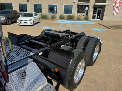 Kenworth T680 truck chassis view showing dual rear axles, fifth-wheel mount, and diamond-plate toolbox; front of a dealership building in background.