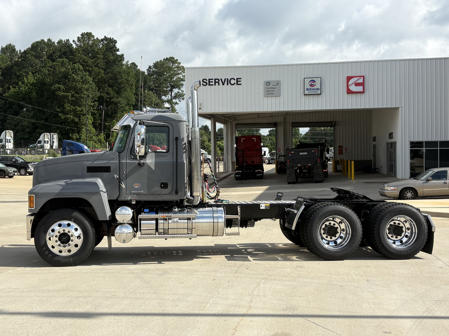 2027 Mack PI64T truck chassis with cab and long wheelbase, parked in front of service center. Visible text includes 'PI64T' on cab and various service signs on building.