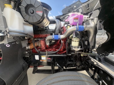 Engine bay of a 2027 Mack PI64T conventional daycab showing red engine block, black hoses, coolant reservoir, and purple coolant overflow cap.