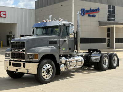 Mack PI64T truck in showroom lot with Bruckners building in background; front grille with Mack logo, chrome bumper, dual fuel tanks, dual rear axles, gray cab, chrome mirrors and exhaust stacks.