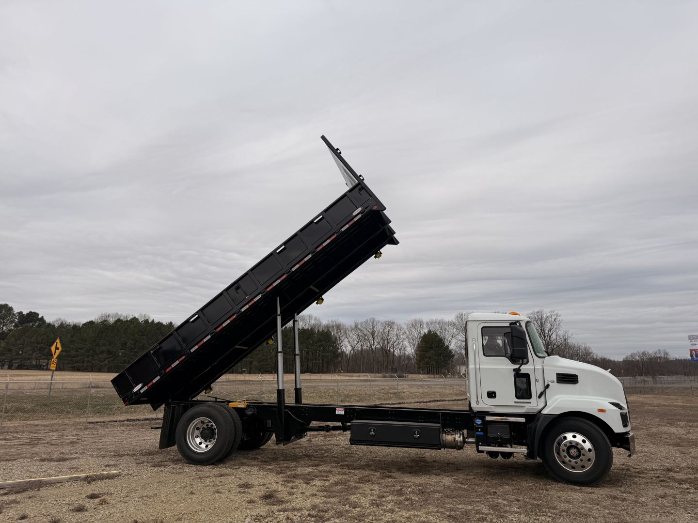 Truck flatbed dump bed in raised position showing white Mack MD7 cab with black tilted dumping bed on a gravel lot.