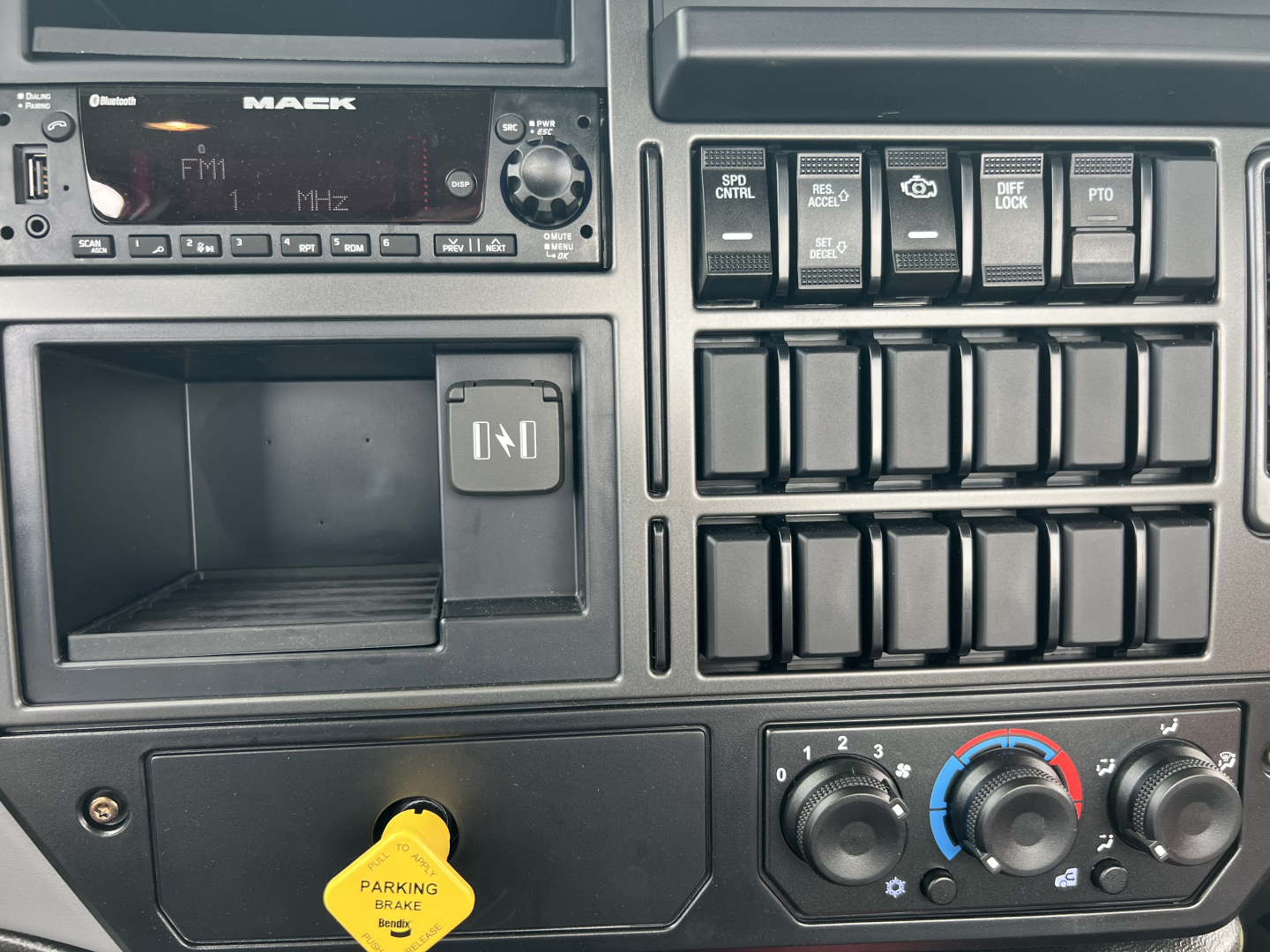 Mack MD7 cabin dashboard showing radio, empty tray, blank switches, and climate controls with yellow parking brake knob visible.