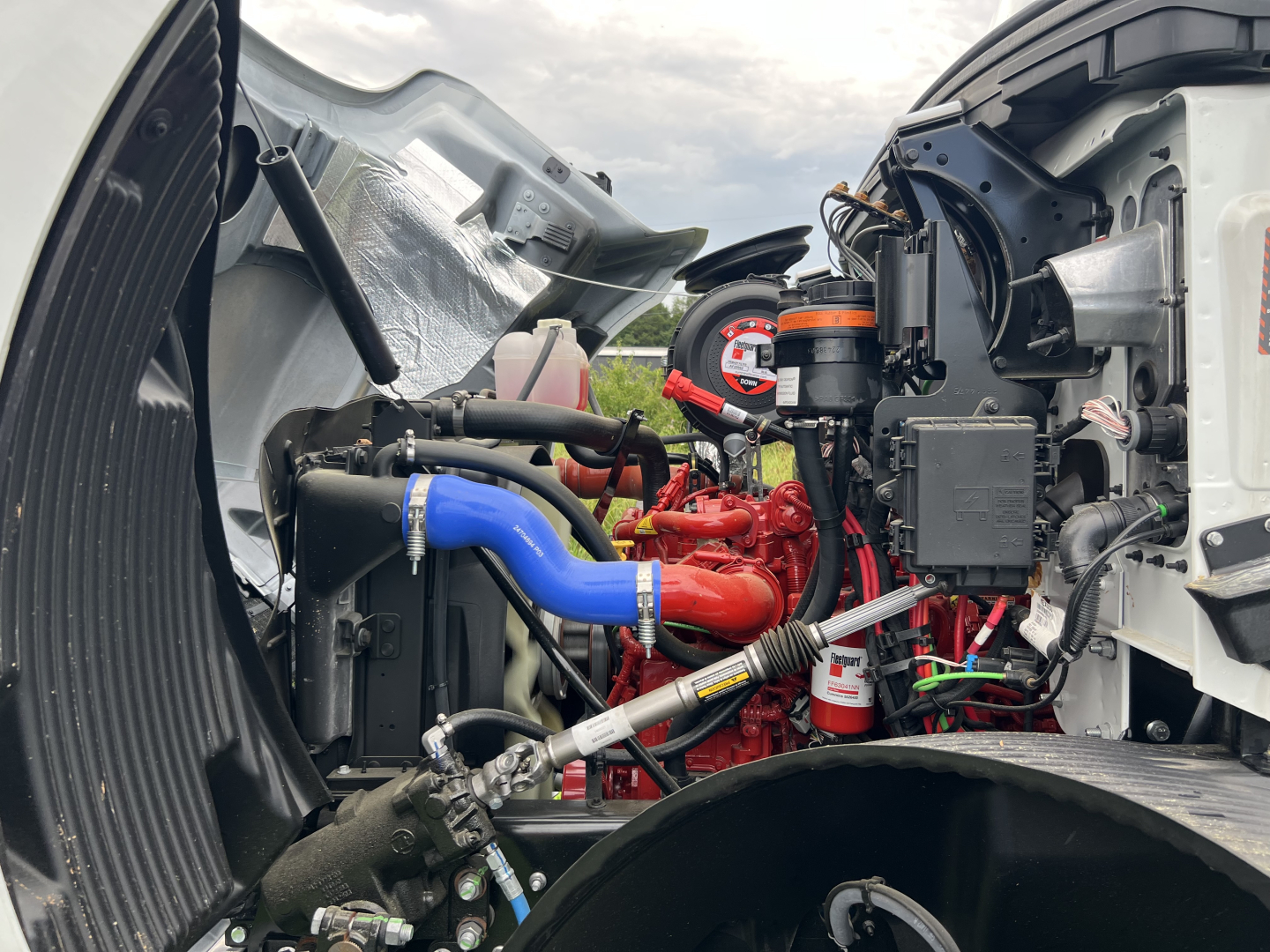 Engine bay of a 2025 Mack MD7 flatbed truck showing blue and red hoses, various connectors and components, intake/air cleaner area in view.