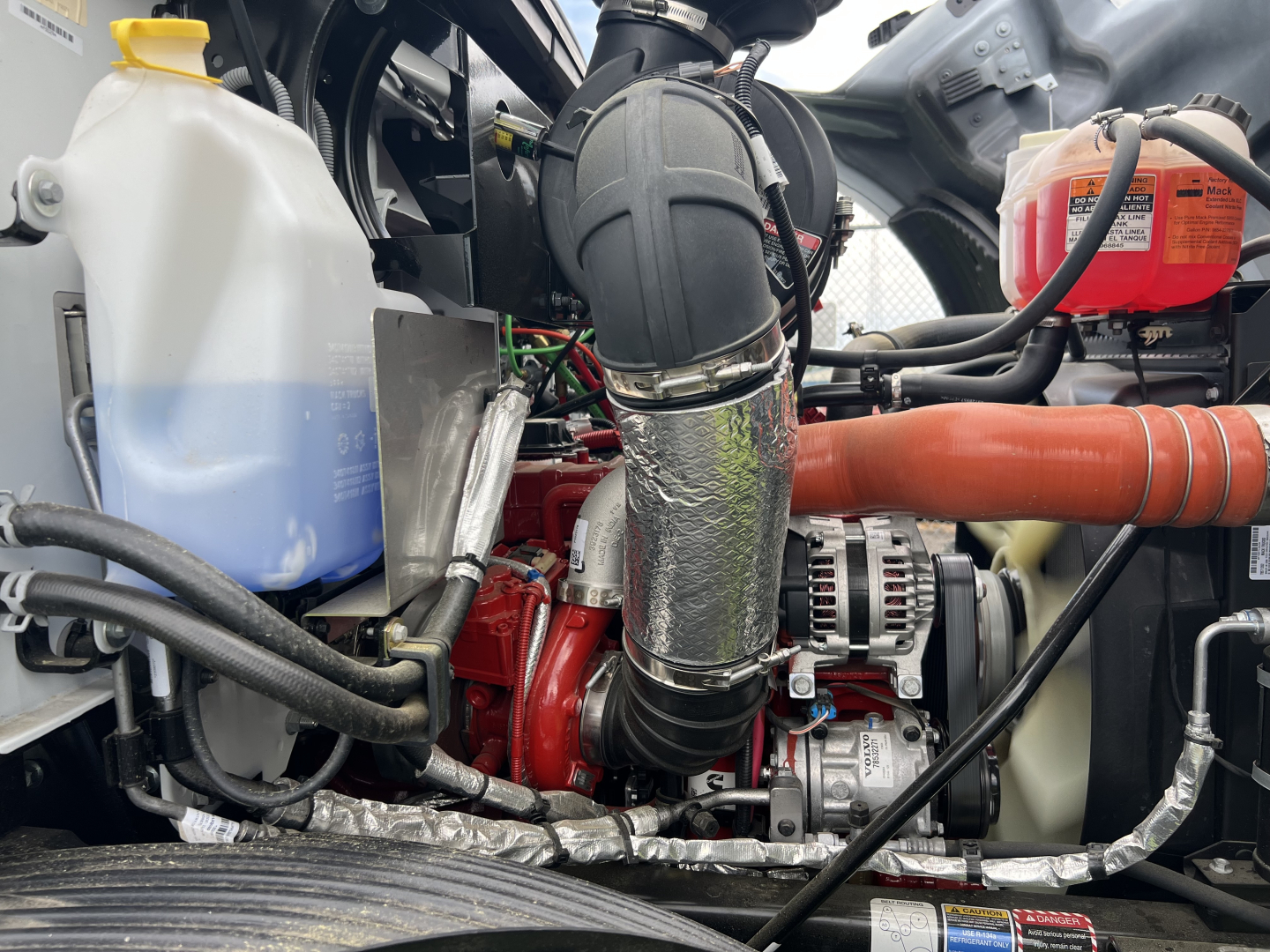 Mack MD7 engine bay showing coolant reservoir, coolant hose, air intake, turbocharger, alternator, and miscellaneous hoses in a flatbed truck. Visible labels include a white and blue coolant container and red coolant bottle.