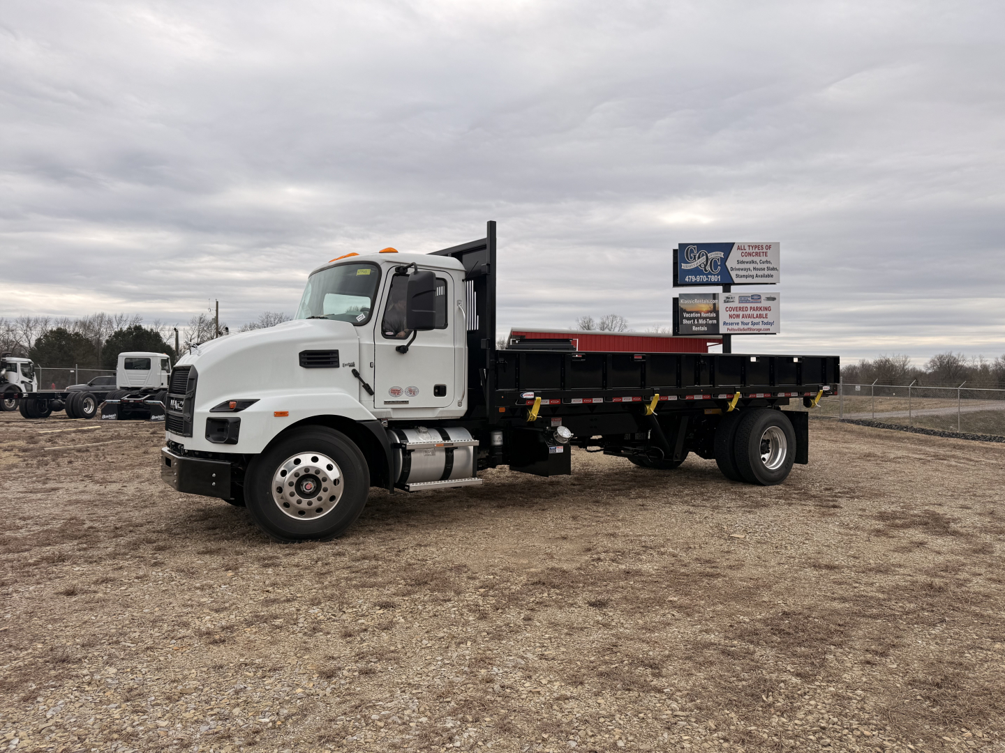 Mack MD7 flatbed truck in a gravel lot showing cab and long flatbed chassis with sign in background