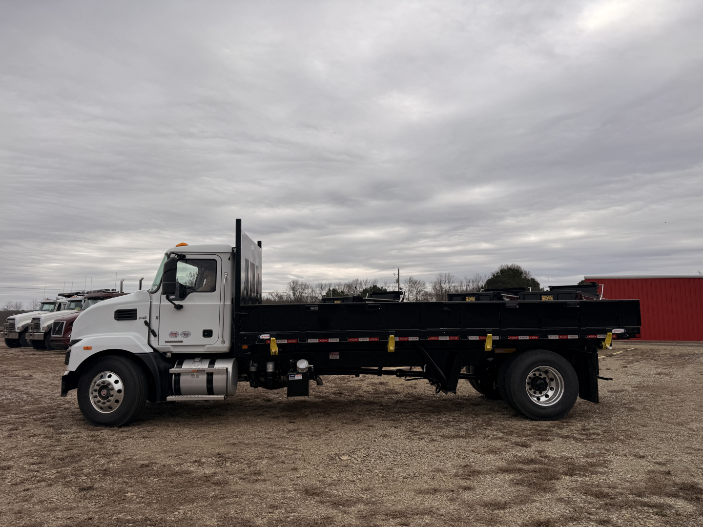Side view of a white cab Mack MD7 flatbed truck with black dump bed, visible tires, and overcast sky.