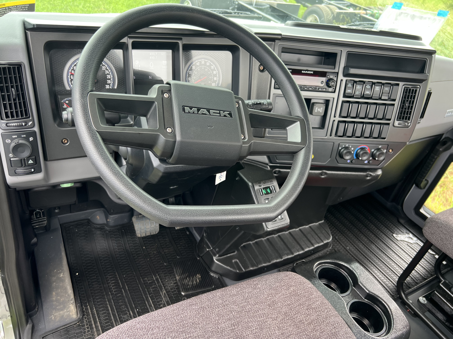 Mack MD7 truck interior showing steering wheel with MACK logo, dashboard gauges, climate controls, and center console; black ribbed floor mats and cup holders.