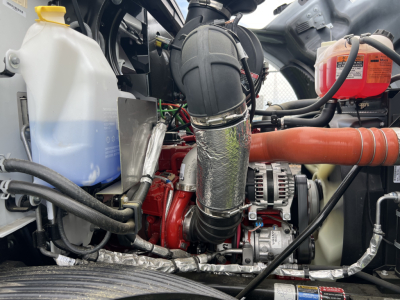 Mack MD7 engine bay showing coolant reservoir, coolant hose, air intake, turbocharger, alternator, and miscellaneous hoses in a flatbed truck. Visible labels include a white and blue coolant container and red coolant bottle.