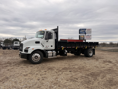 Mack MD7 flatbed truck in a gravel lot showing cab and long flatbed chassis with sign in background