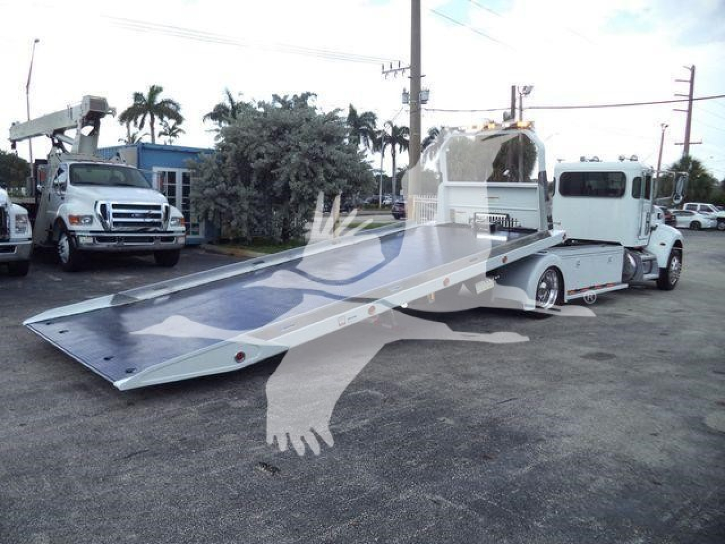 Truck truck bed wrecker-style rollback with a long aluminum ramp extended, Peterbilt 337 cab visible in background, service lot setting.