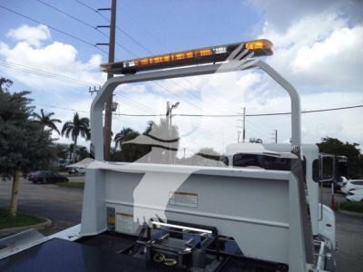 Rear view of a white Peterbilt rollback truck with a safety frame and warning lights on top. Visible text includes small labels on the dump bed and a central hitch mechanism.