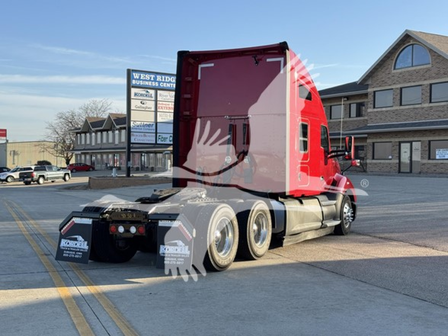 Kenworth T680 sleeper cab tractor viewed from rear-left, red cab, dual rear axles, third axle visible, on lot with dealership signs