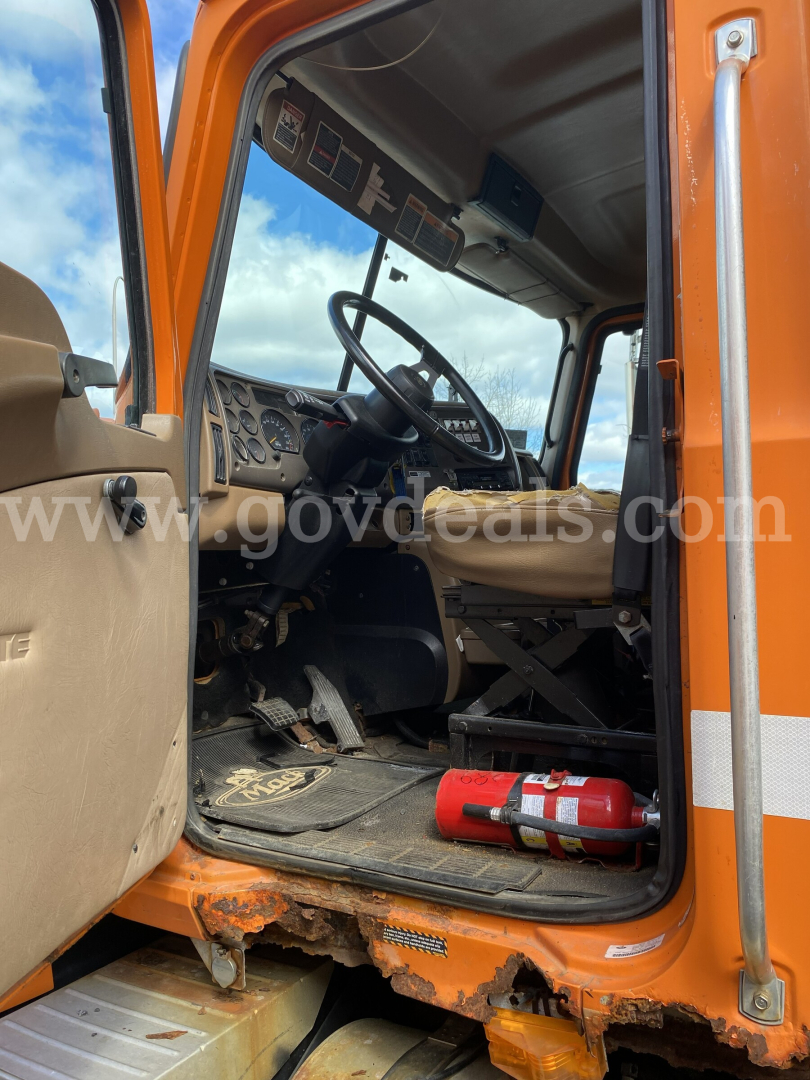 Cab interior of a 2004 Mack CV713 Granite dump truck with orange exterior, showing steering wheel, dashboard, and driver seat.