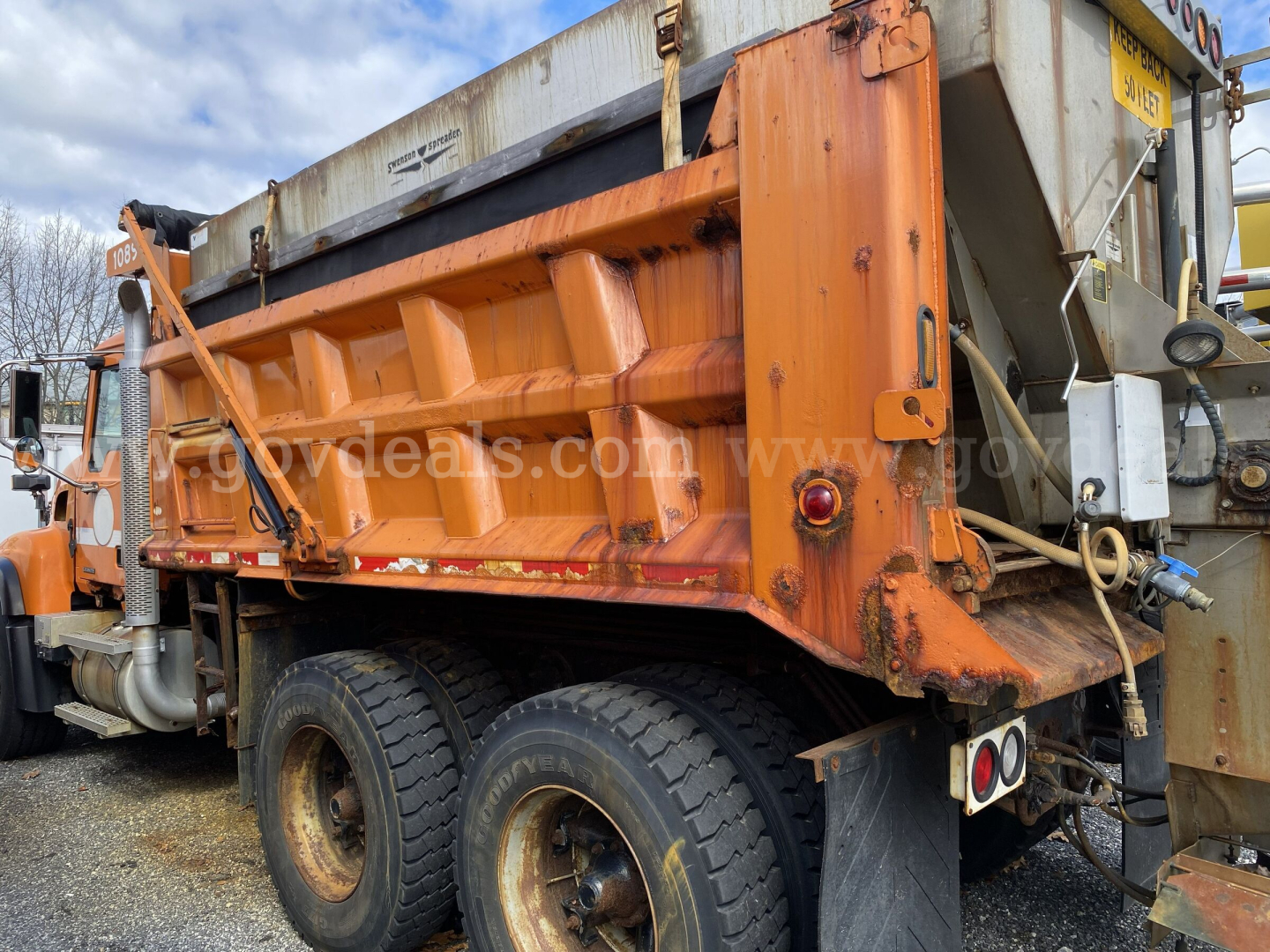 Side view of a 2004 Mack CV713 Granite dump truck, orange bed, dual rear wheels, with various hydraulic components and a metal hopper. Visible text includes stock/ID markings on the truck and site watermark.