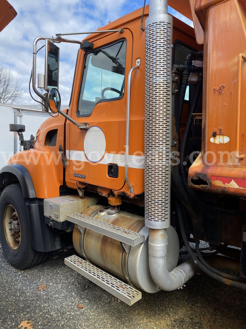 Truck cab side view of a orange 2004 Mack CV713 Granite dump truck showing chrome exhaust stack, side mirror, steps, and fuel tank.