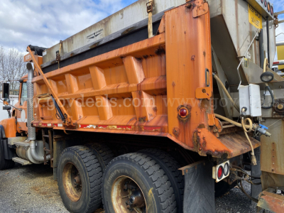 Side view of a 2004 Mack CV713 Granite dump truck, orange bed, dual rear wheels, with various hydraulic components and a metal hopper. Visible text includes stock/ID markings on the truck and site watermark.