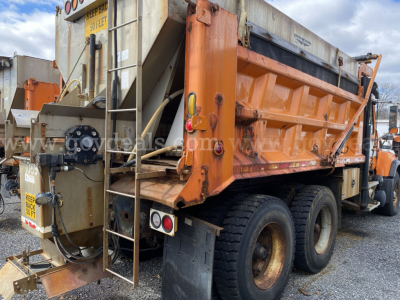 Dump truck rear view showing orange steel dump bed, dual rear axles, and ladder with safety cage. Visible are rear lights, hydraulic hoses, and metal frame components.