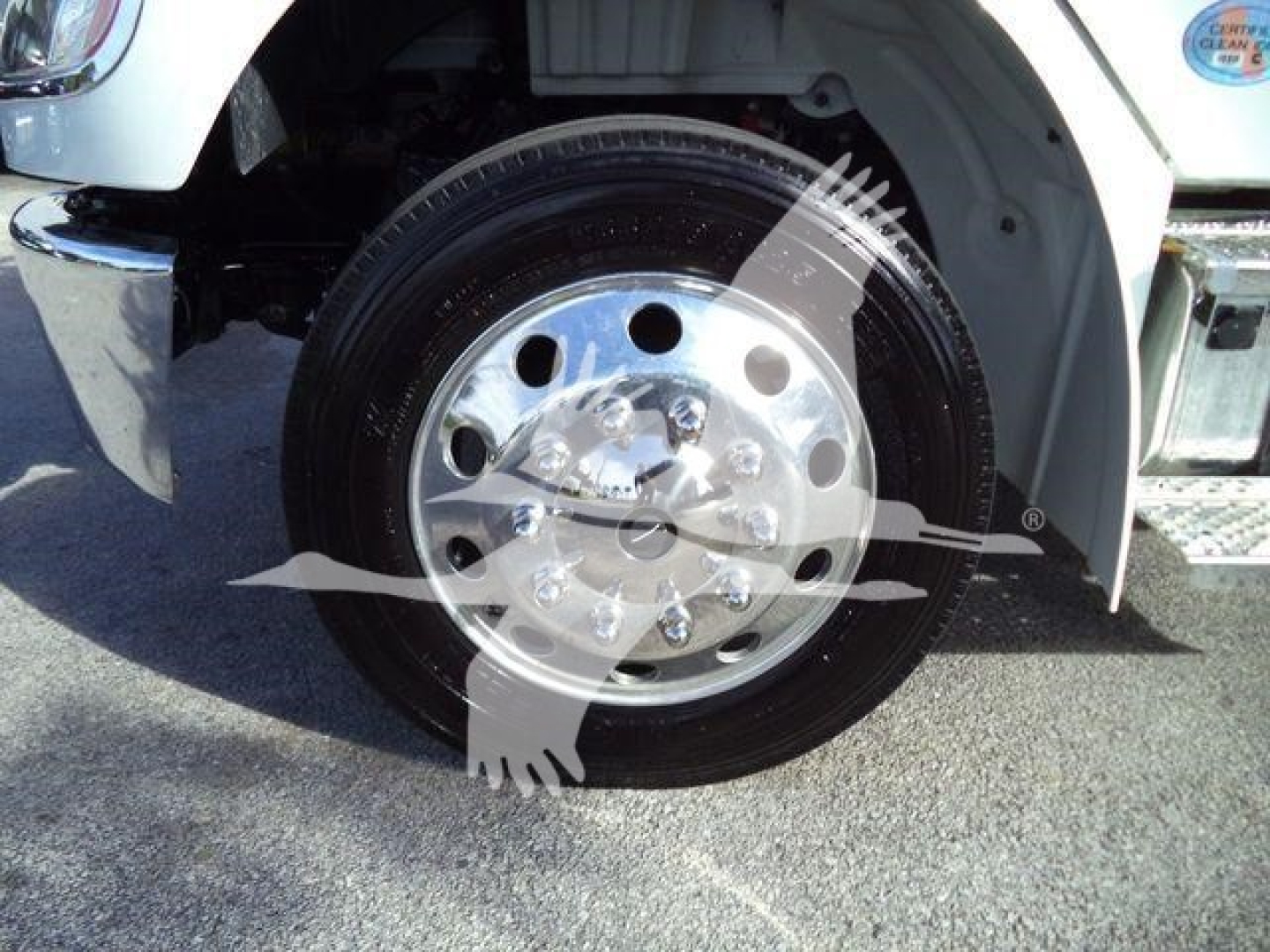 Front wheel of a Freightliner M2 106 Plus truck showing chrome alloy wheel and tire, with part of the white fender and side chrome bumper.