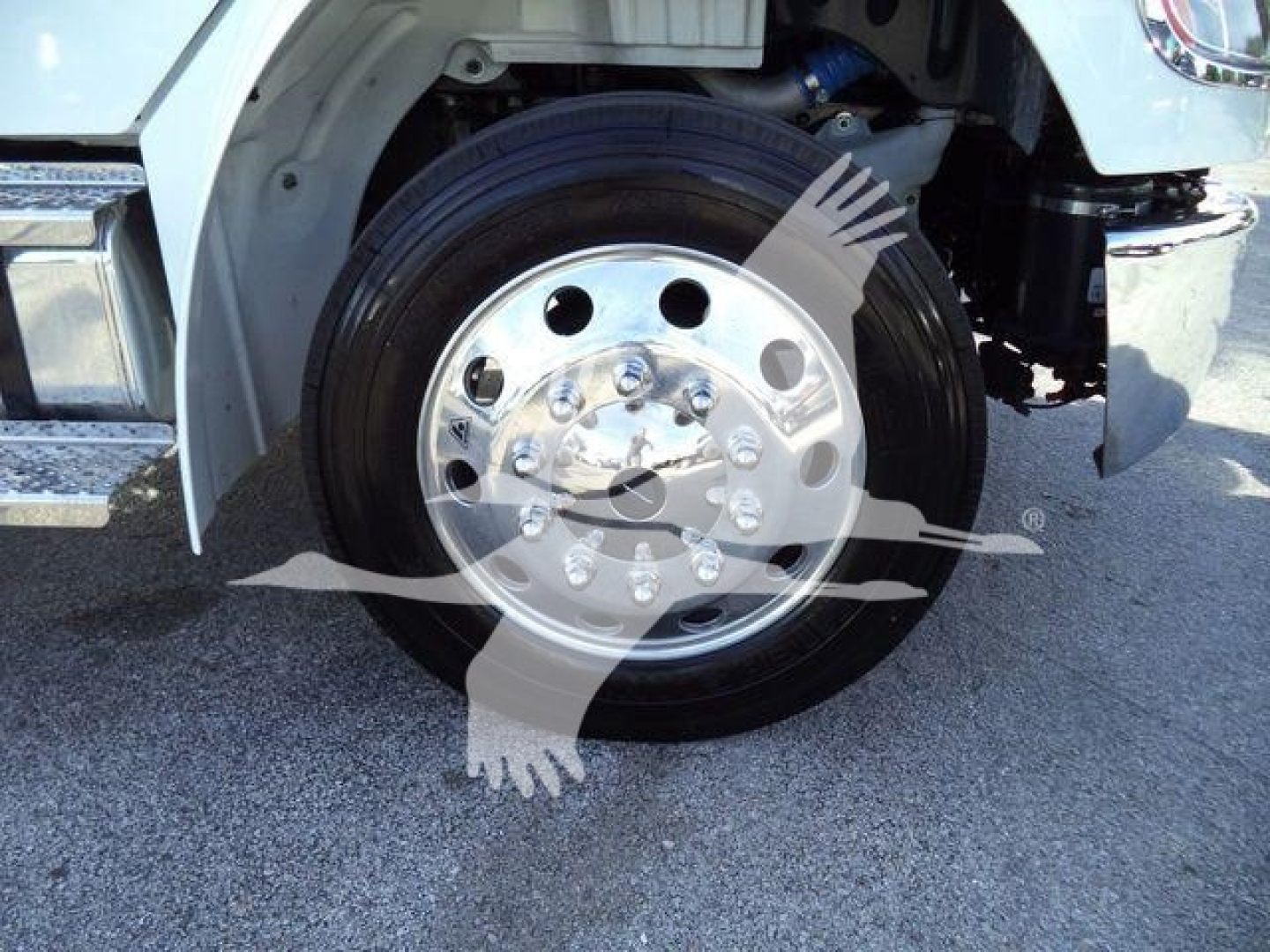 Wheel and front axle of a Freightliner M2 106 Plus truck showing polished aluminum hub with lug nuts and black tire; partial front fender and bumper visible.