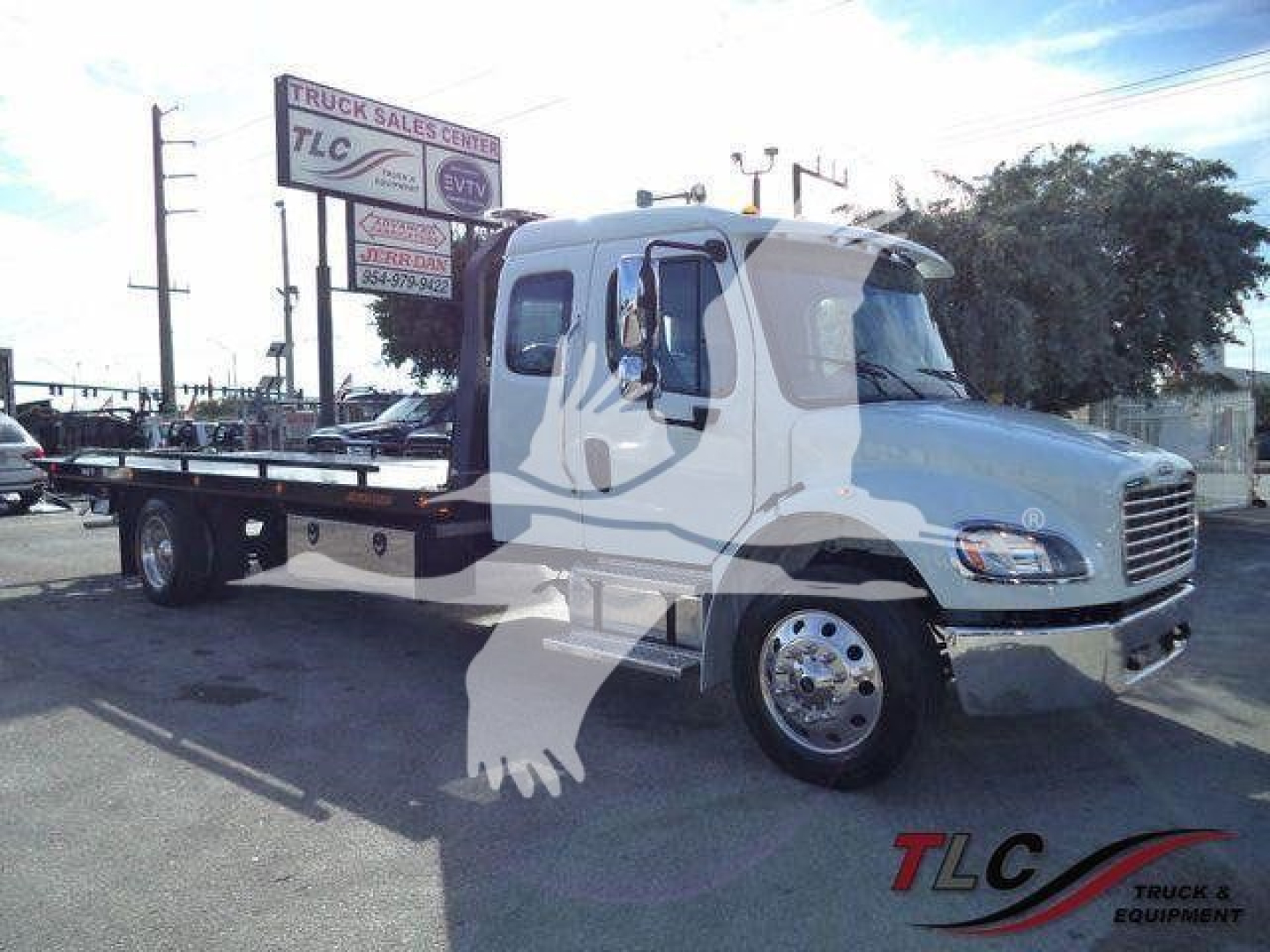 Freightliner M2 106 Plus rollback truck viewed from side, white cab with flatbed, chrome wheels, dealership signage in background.