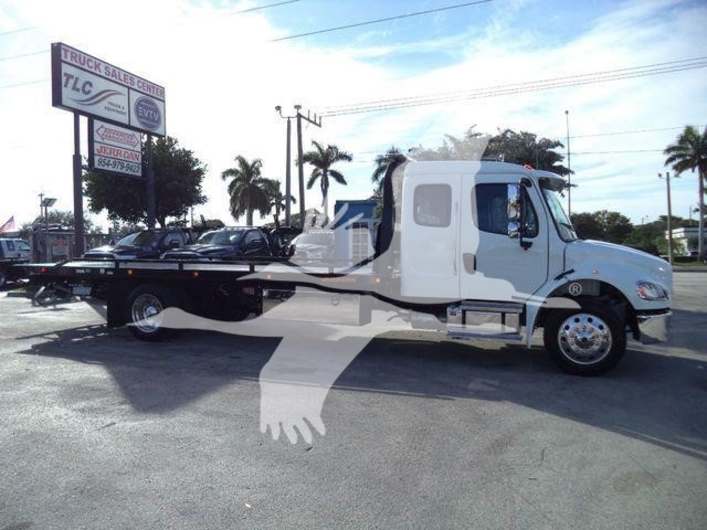 Truck with a rollback wrecker body, Freightliner M2 106 Plus style cab, light blue and white color, stock sign in background, open lot.