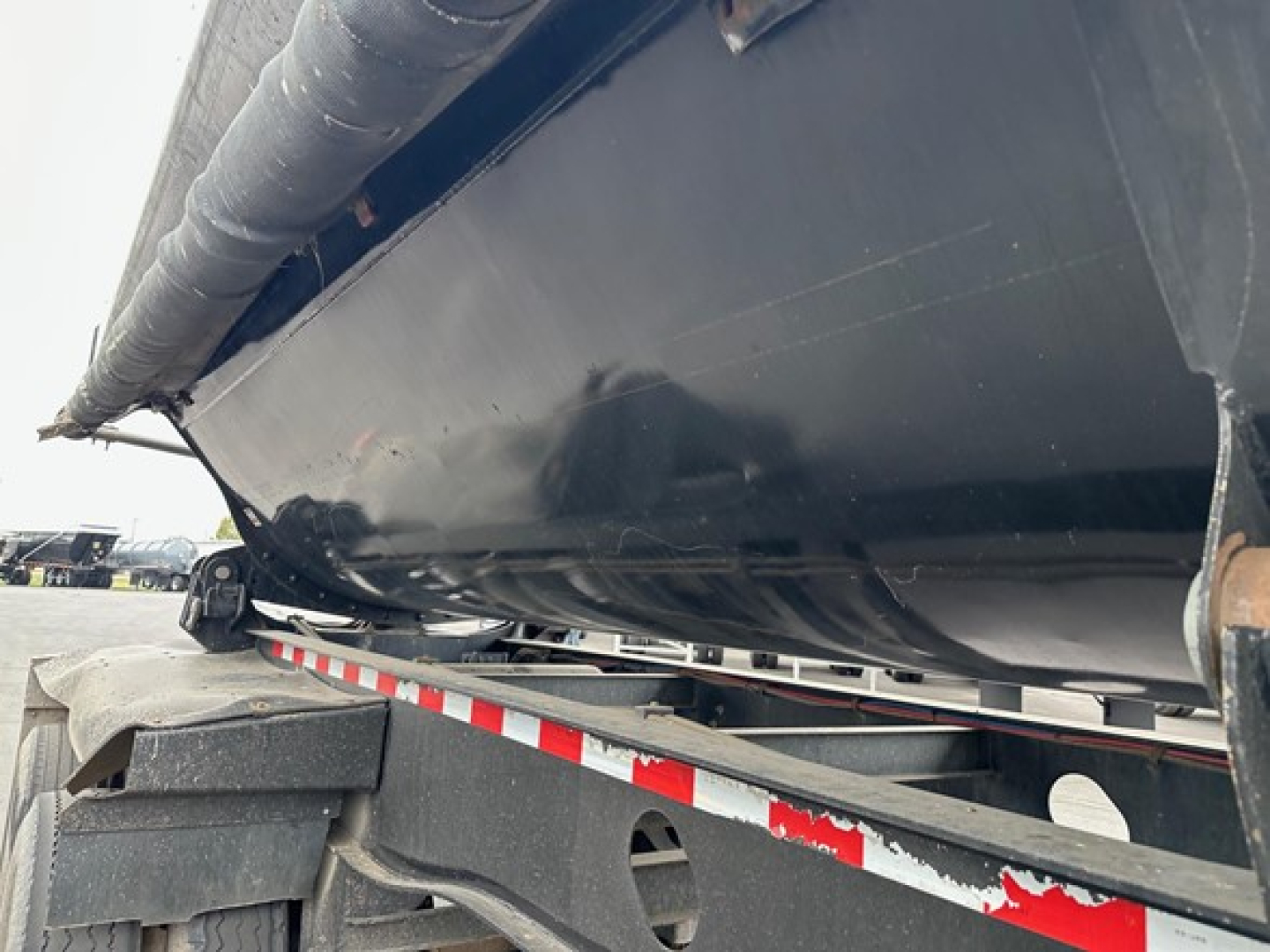 Dump trailer underside showing side dump mechanism and structural frame with black paint and reflective red-and-white safety tape label.