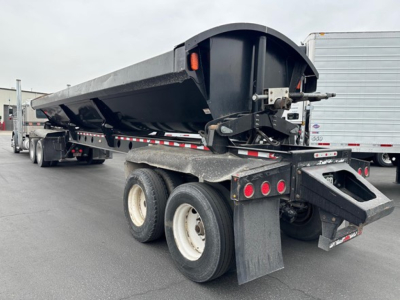 Dump trailer showing Jet side dump with hydraulic hoist and black steel bed. Dual rear axles, white wheels, mudflaps, and rear bumper with lights visible.