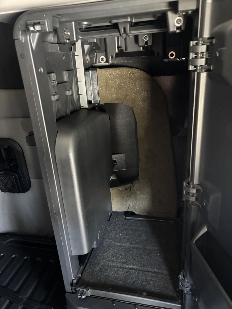 Cab interior area showing HVAC or storage compartment under dashboard with plastic trim and wiring harness; empty small cavity with dark carpeting.