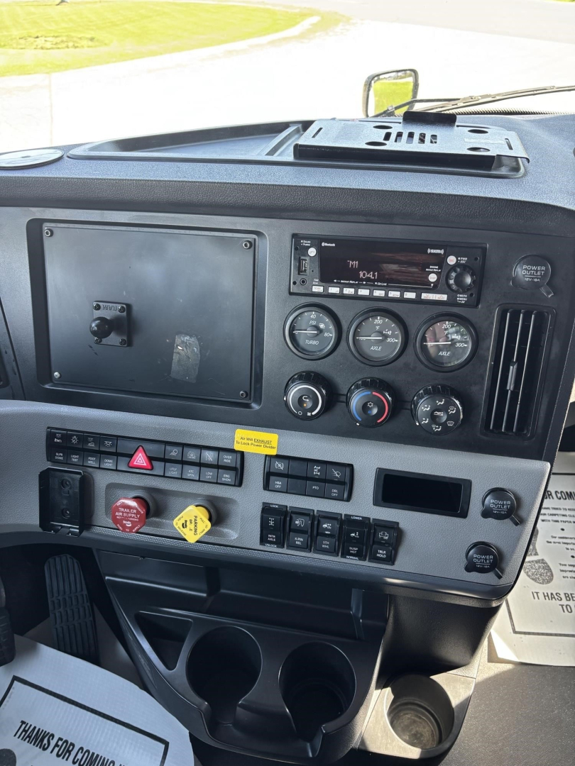 Truck cab dashboard of a 2021 Freightliner Cascadia 126 showing steering column area, instrument cluster, climate controls, switches, and storage compartments.