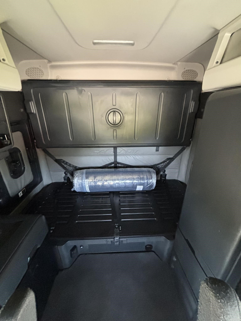 Truck sleeper cab interior showing storage panel and horizontal cylindrical air tank under panel in Freightliner Cascadia engine bay area.