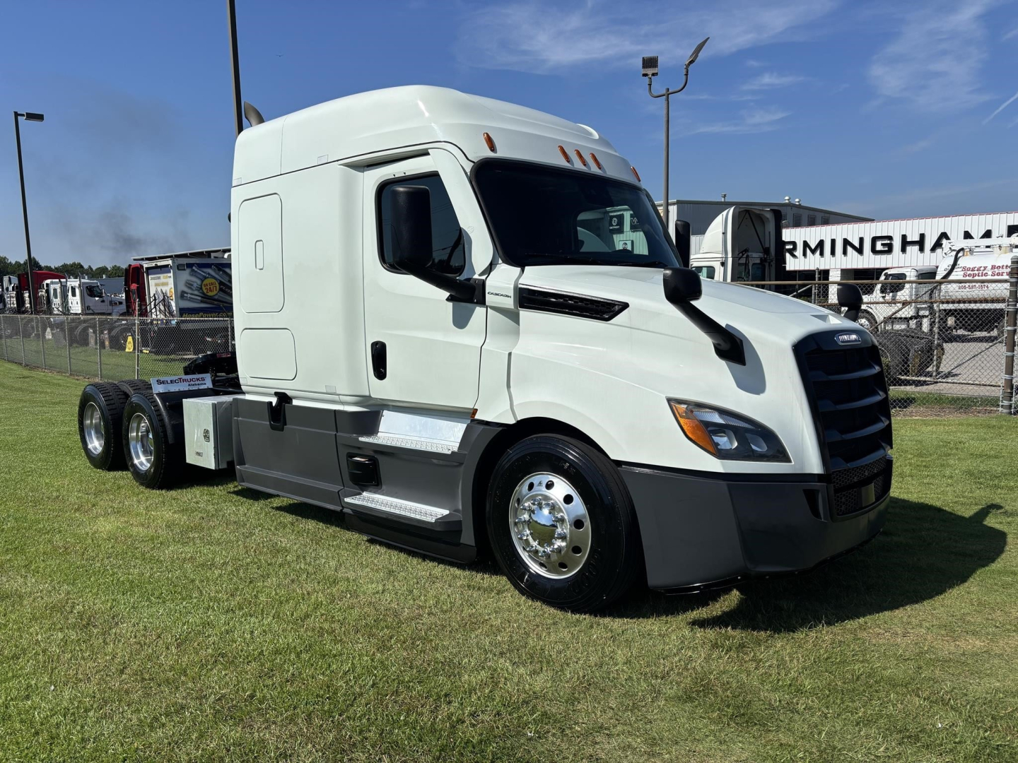 Freightliner Cascadia 126 truck in a grassy lot with white cab, black bumper, chrome wheels. Visible text on truck: none clearly legible; a sign in background reads ‘Birmingham’ and there are no model decals on cab visible.
