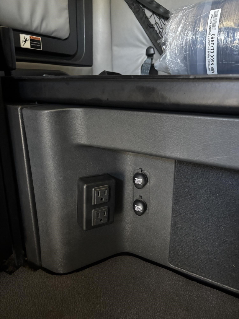 Cab area of a Freightliner Cascadia showing a dual USB/aux panel and two round buttons near a storage compartment.