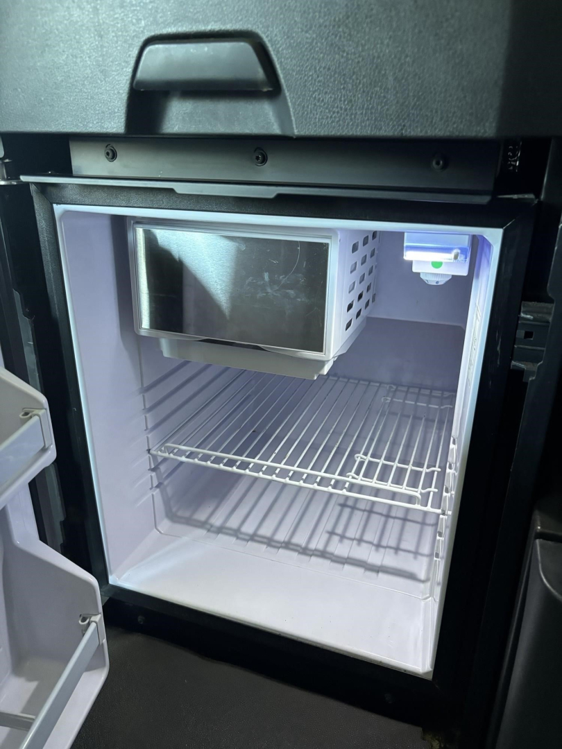 Mini fridge/freezer mounted inside a Freightliner Cascadia cab, open door showing an empty freezer with wire rack shelf and a small microwave on top inside the unit.