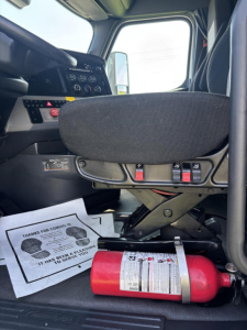 Truck cab interior showing driver seat base and control panel with switches, fire extinguisher on floor, and paperwork on the step.